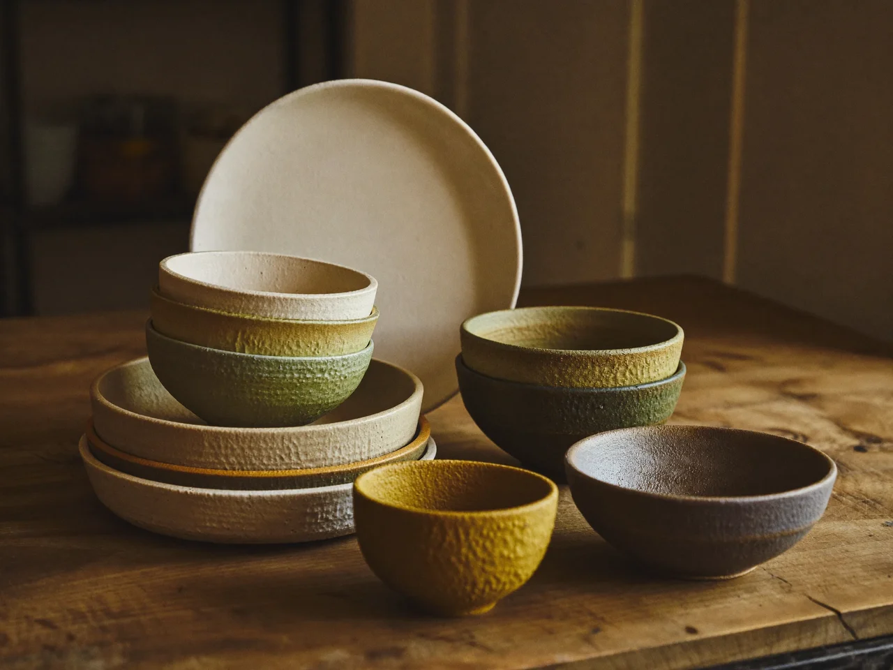 Pila di piatti e ciotole in ceramica opaca dai toni terrosi su piano in legno